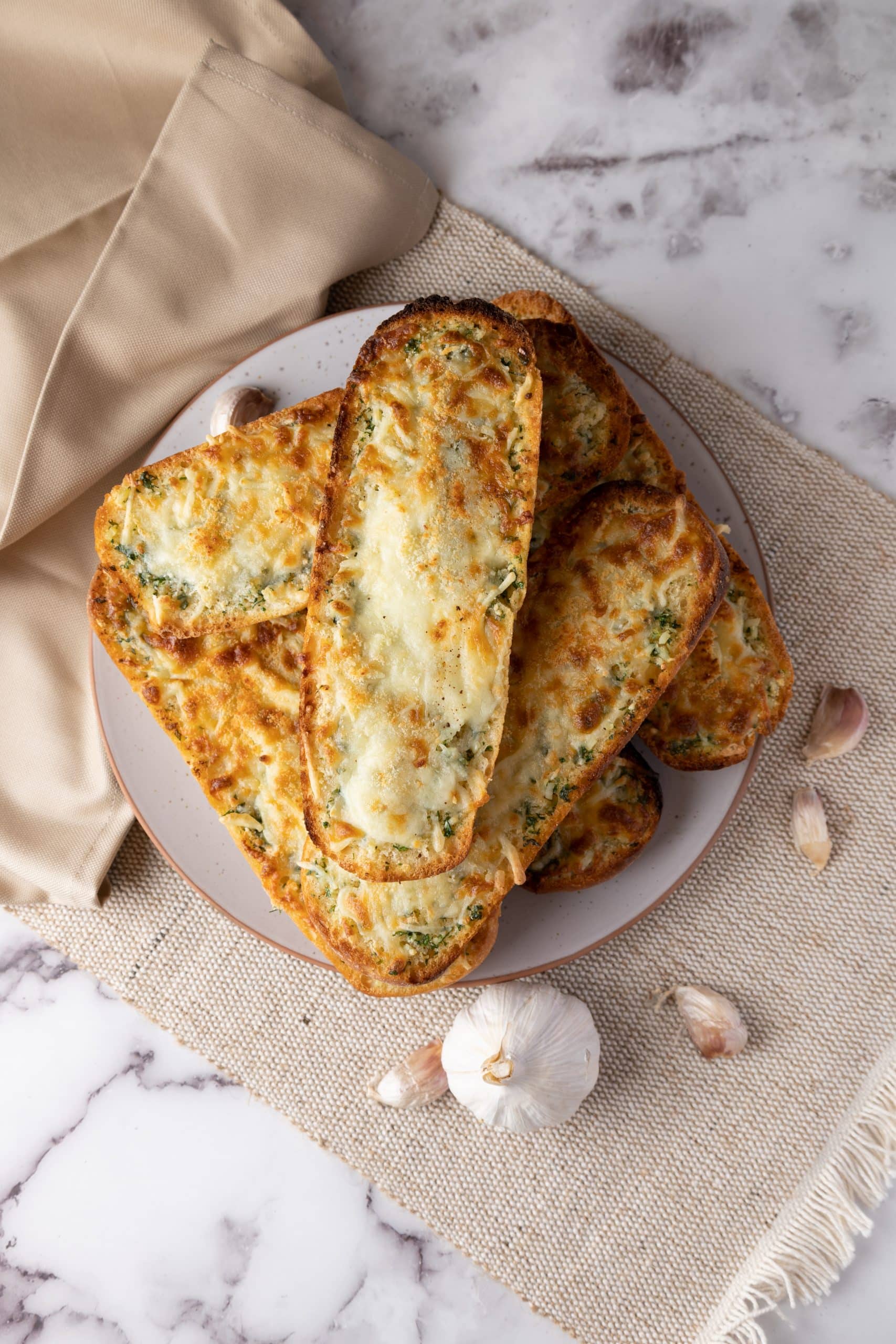 Halved loaf baked with melted mozzarella, Parmesan and parsley on a plate beside garlic bulbs