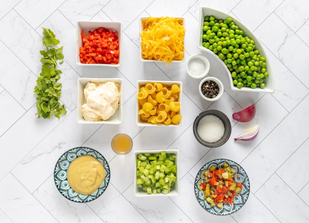 An array of fresh ingredients ready to be transformed into the best macaroni salad recipe - elbow noodles, cheddar cheese, red onion, celery, red bell pepper, peas, mayonnaise, cider vinegar, fresh dill, sugar, sweet pickle relish, Dijon mustard, salt, and pepper.