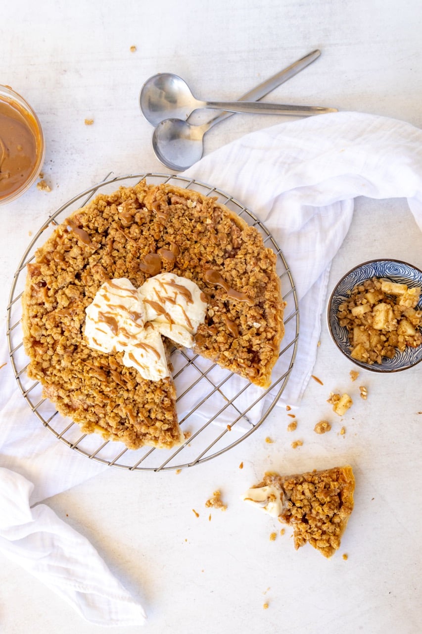 Round crumble topped tart on a wire rack with whipped cream and a drizzle of caramel
