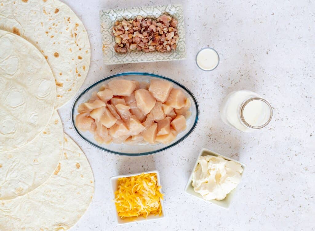 A flat lay of ingredients for chicken pinwheels, featuring cream cheese, ranch seasoning, cheddar, tortillas, and cooked bacon.