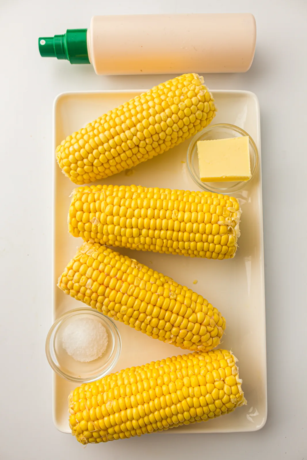 Ingredients for air fryer corn on the cob: four husked ears, butter, salt and oil spray on a tray
