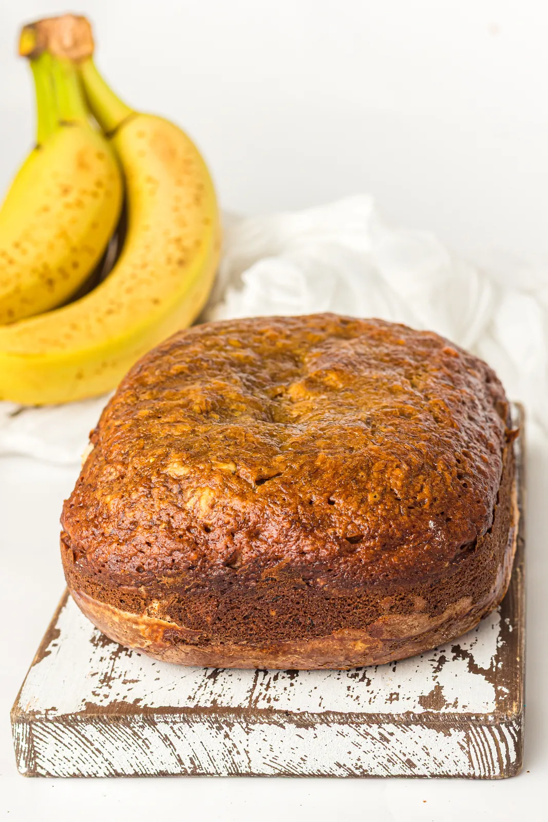 A dark golden banana loaf on a rustic white board with two ripe bananas behind it