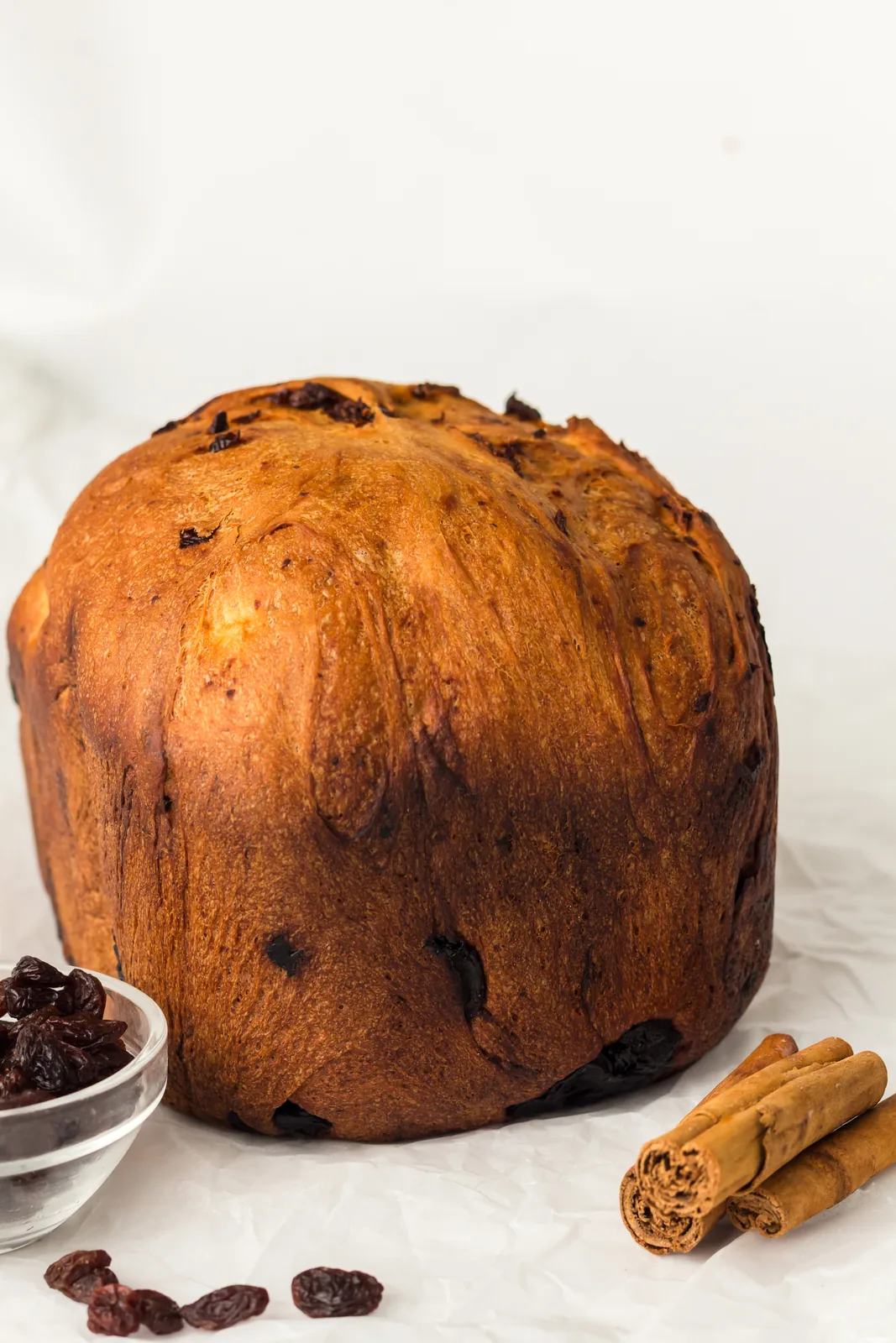 A round topped, deep brown fruit loaf beside a small bowl of raisins and cinnamon sticks