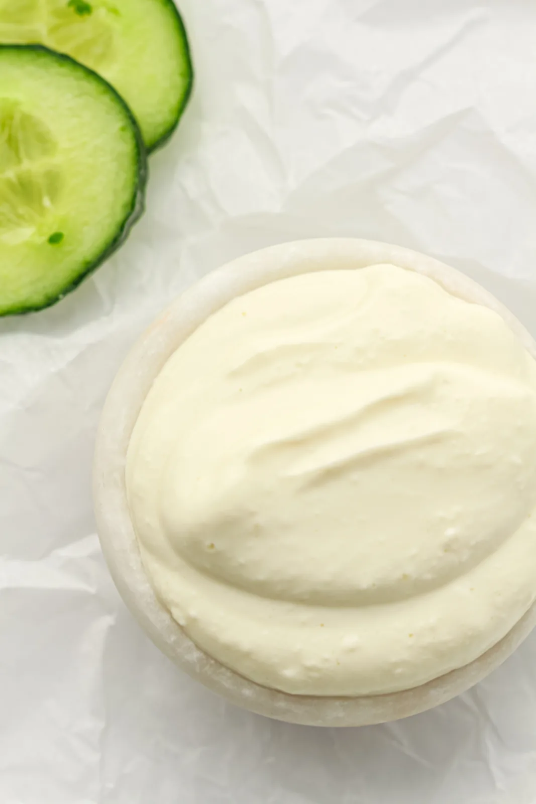 A bowl of homemade cream cheese on parchment with cucumber slices alongside
