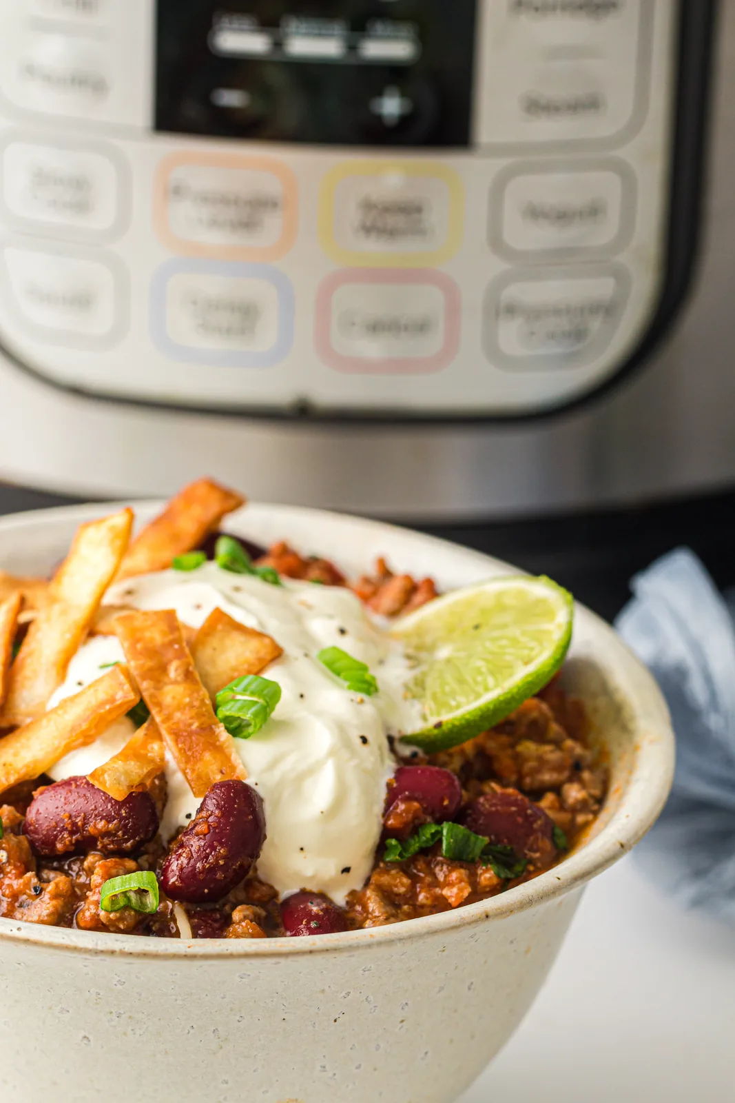 Instant pot chili in a bowl with sour cream, lime and tortilla strips, pressure cooker behind