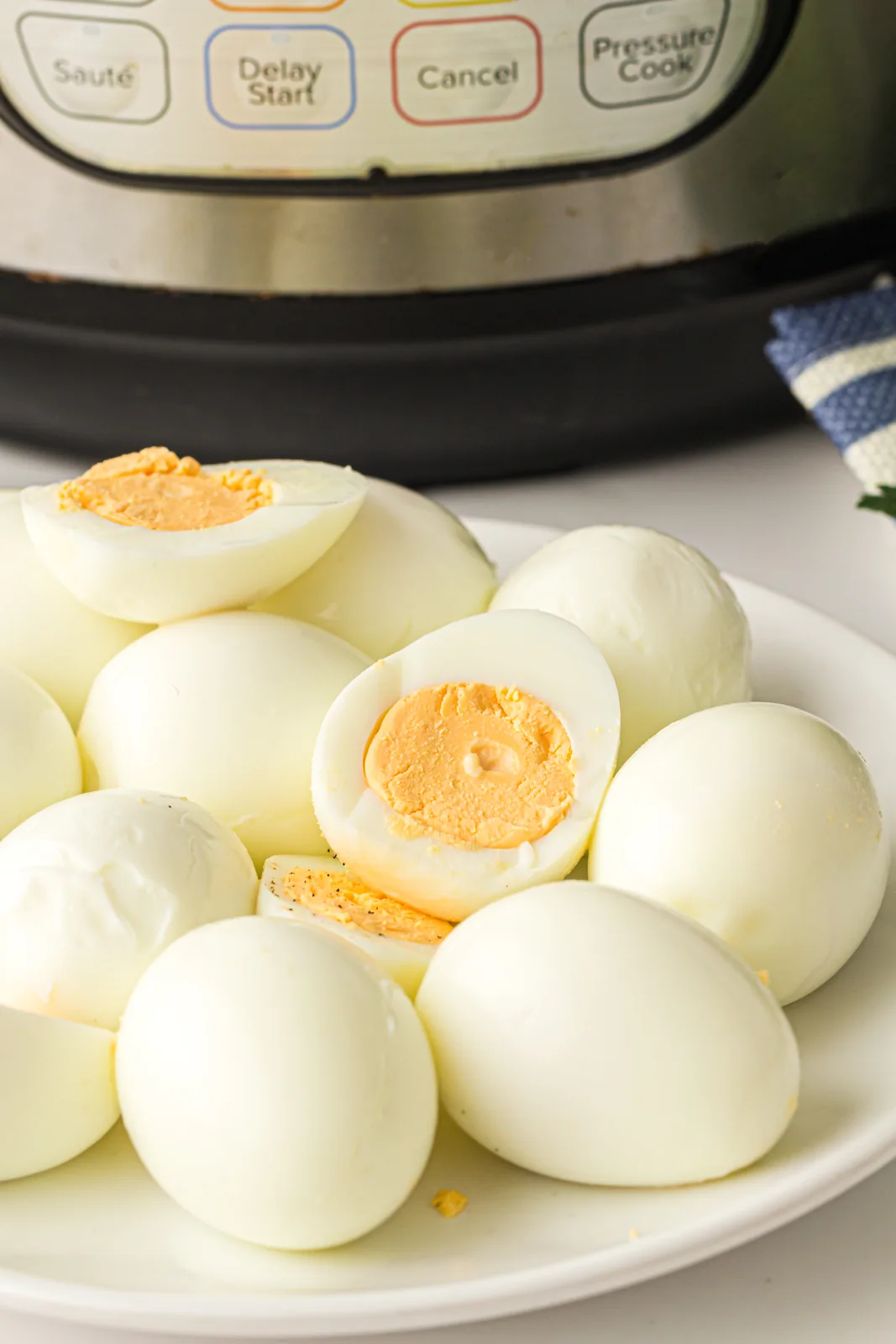 Instant pot hard boiled eggs peeled on a plate, one halved to show the yolk, pressure cooker behind