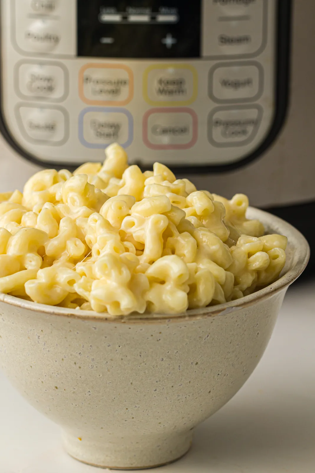 A rustic bowl piled high with creamy yellow elbows, pressure cooker in the background