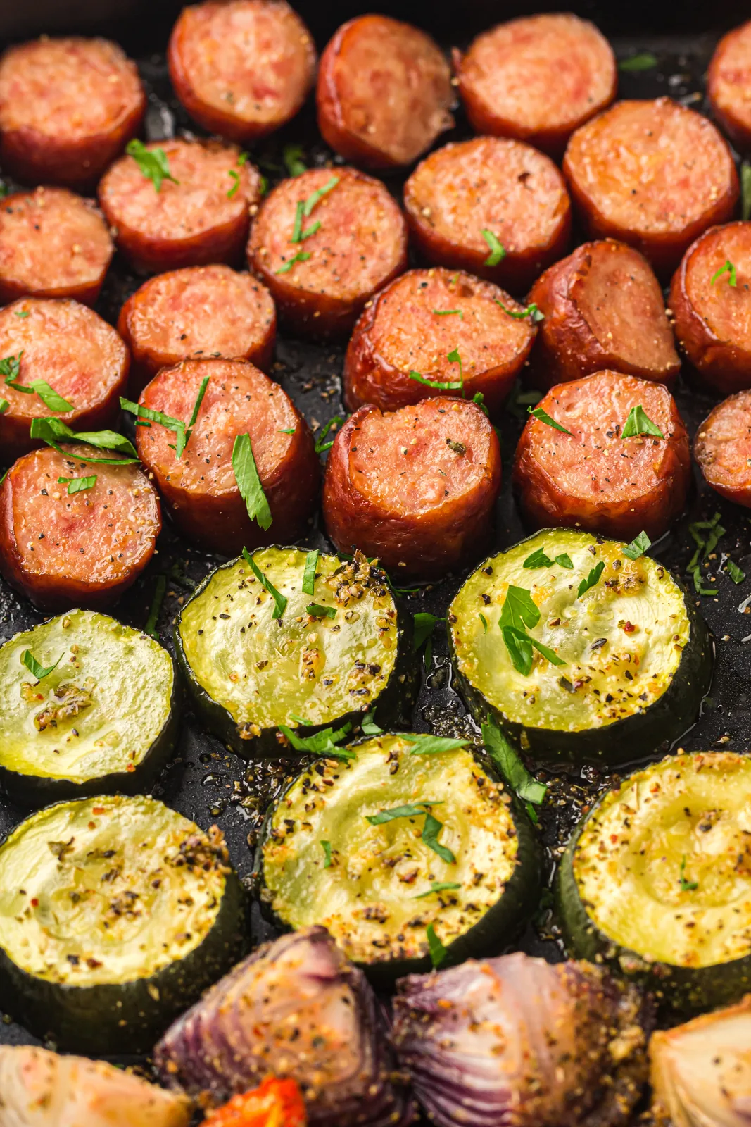 Browned smoked sausage rounds, courgette coins and red onion roasted on a dark tray