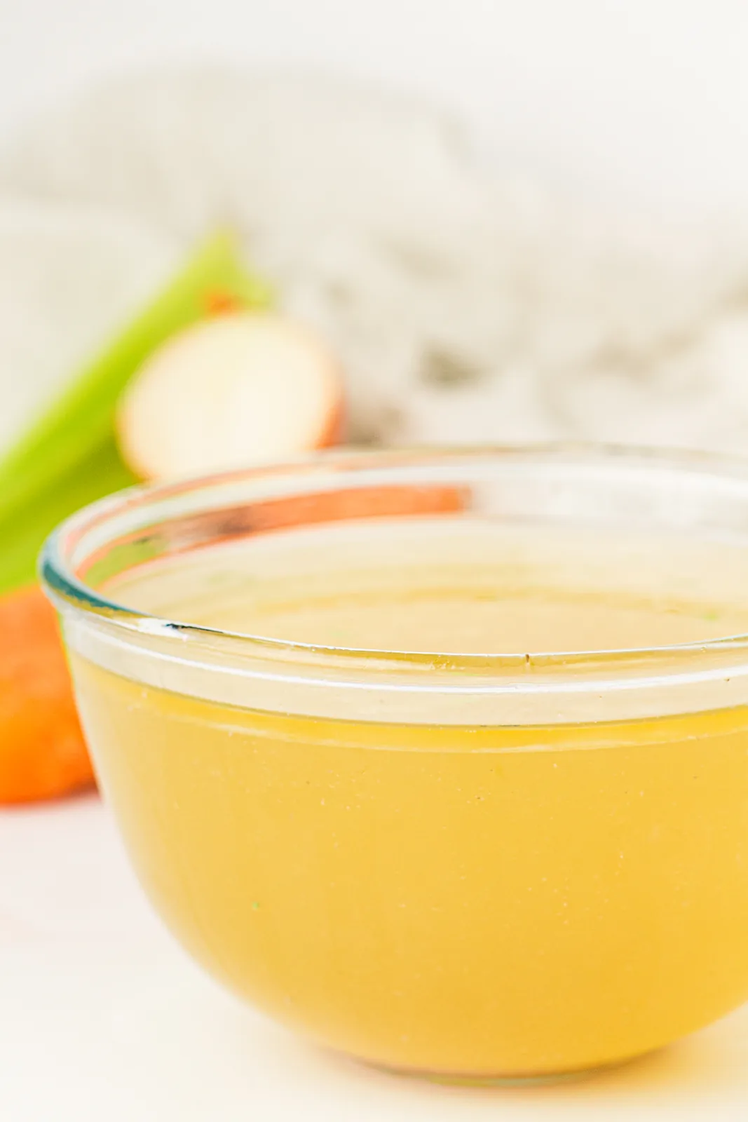 A glass bowl of clear slow cooker chicken stock with carrots and celery behind