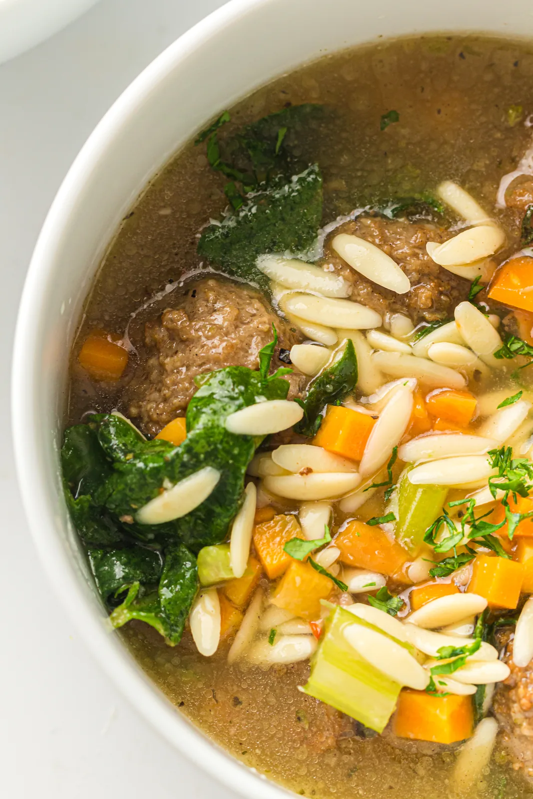Brothy soup with small meatballs, orzo, diced carrot, celery and spinach in a white bowl