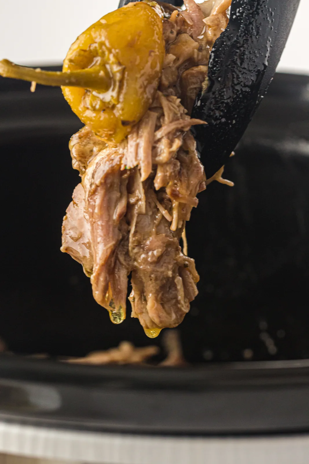 A spatula lifting buttery shreds and a yellow pickled pepper from the crock