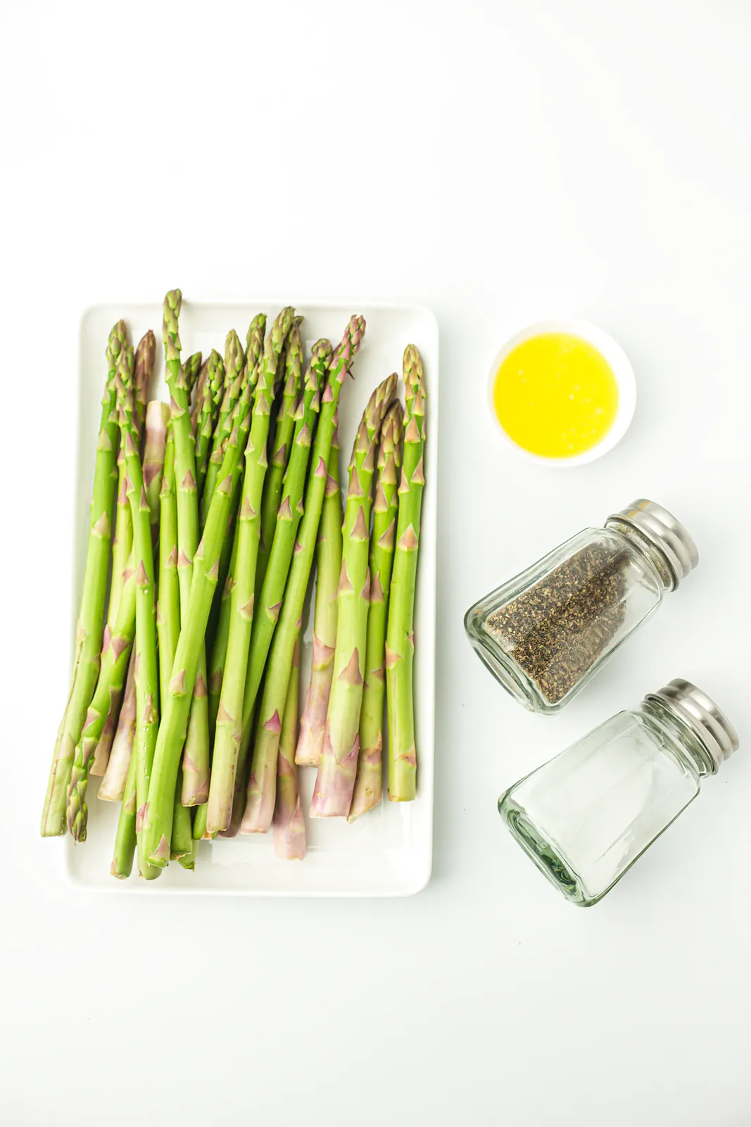 Ingredients for air fryer asparagus: raw spears on a white tray with oil, salt and pepper
