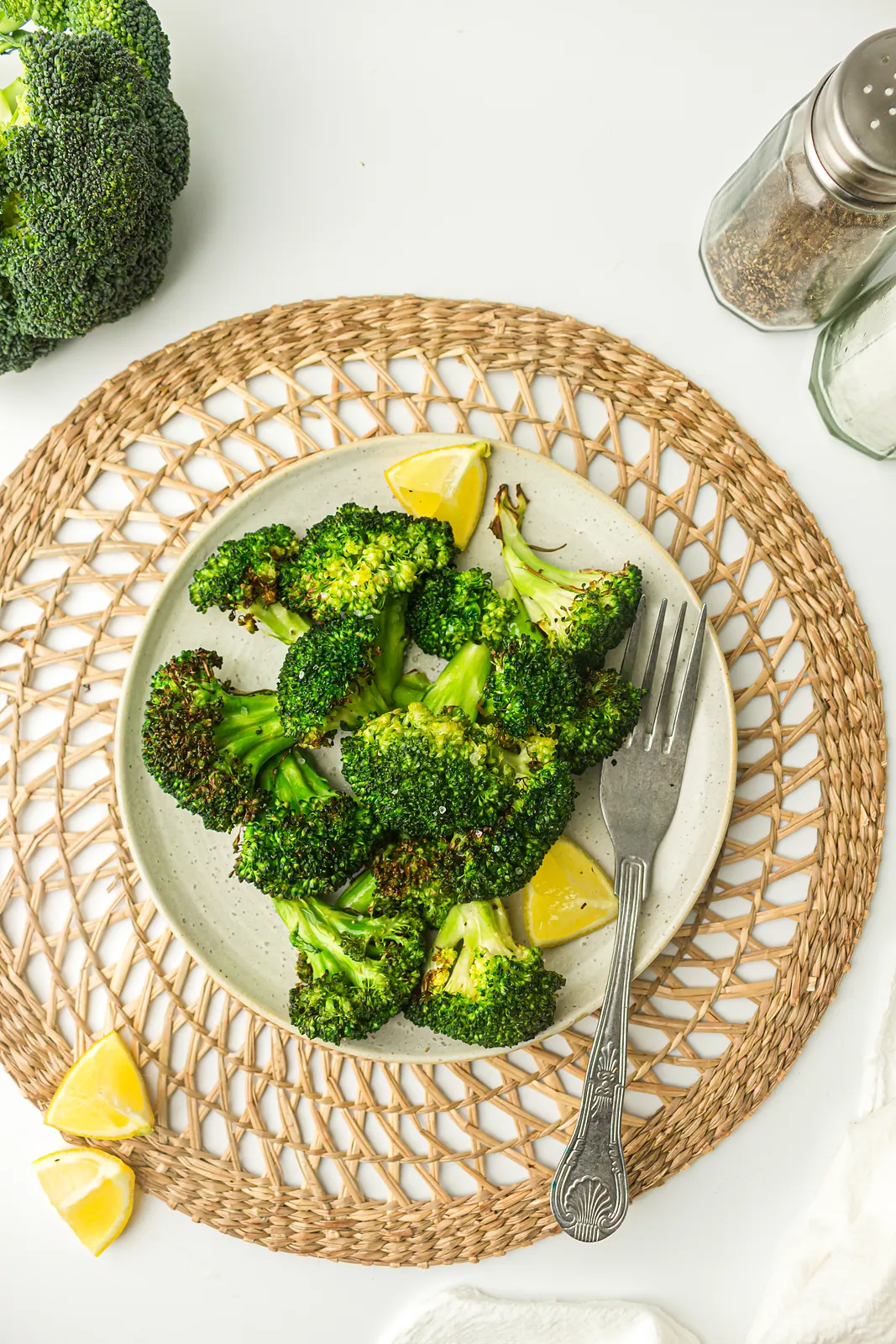 Air fryer broccoli on a plate with lemon wedges and a fork on a woven placemat