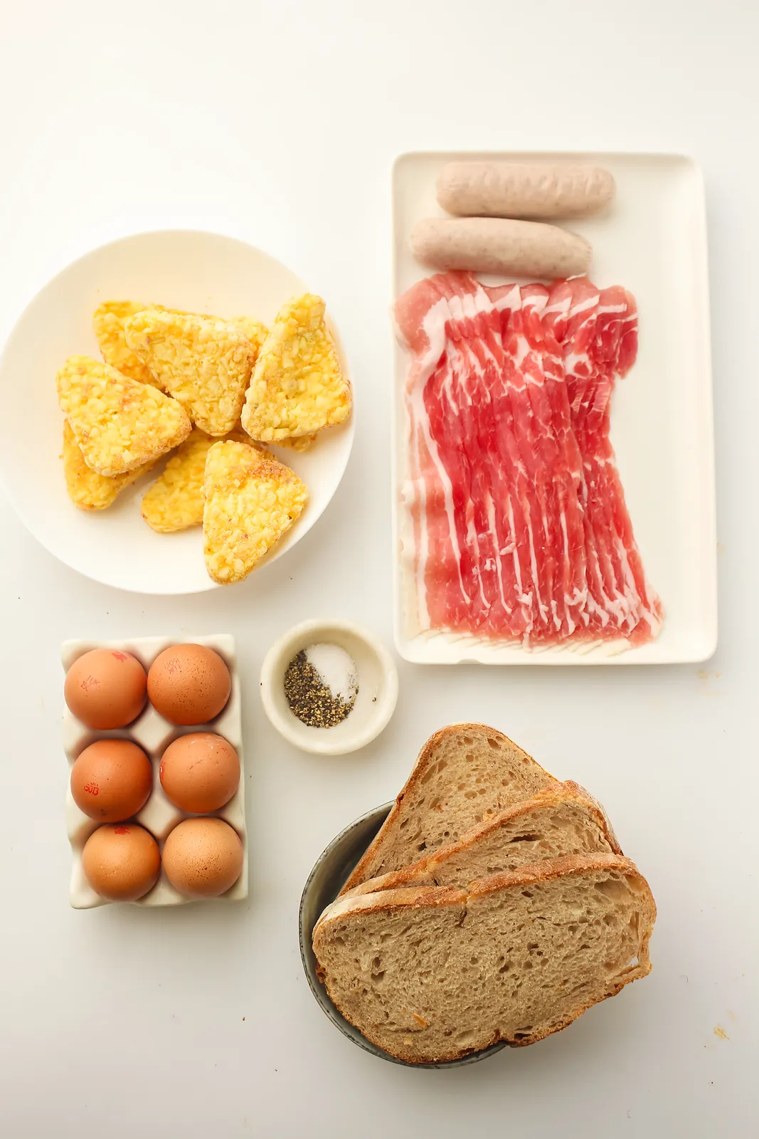 Ingredients for a Blackstone breakfast: raw bacon, pork sausages, frozen hash brown triangles, eggs, pepper and sliced bread