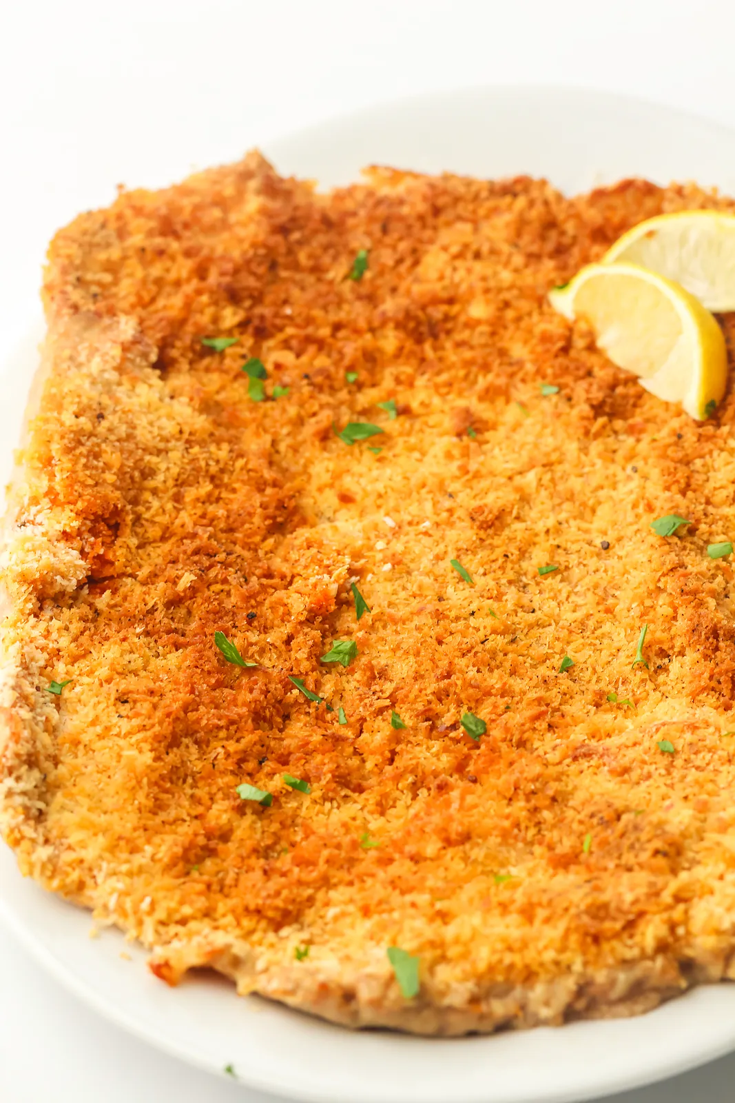 Giant chicken schnitzel on a white plate with lemon wedges and chopped parsley