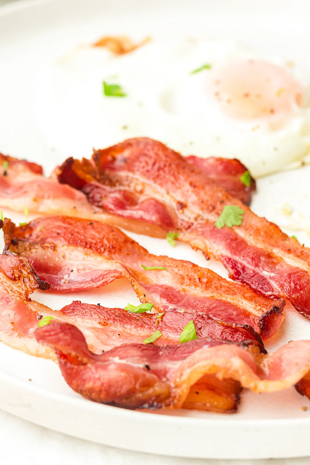 Close up of glossy, crisp rashers on a white plate with a fried egg behind