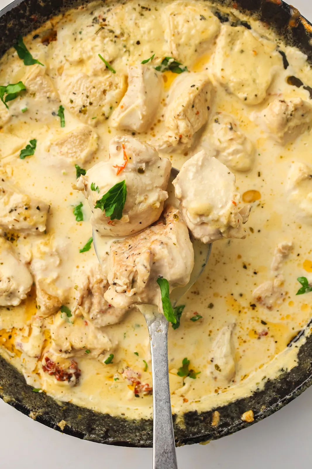 A spoon lifting tender pieces from a creamy Parmesan sauce in a cast iron pan
