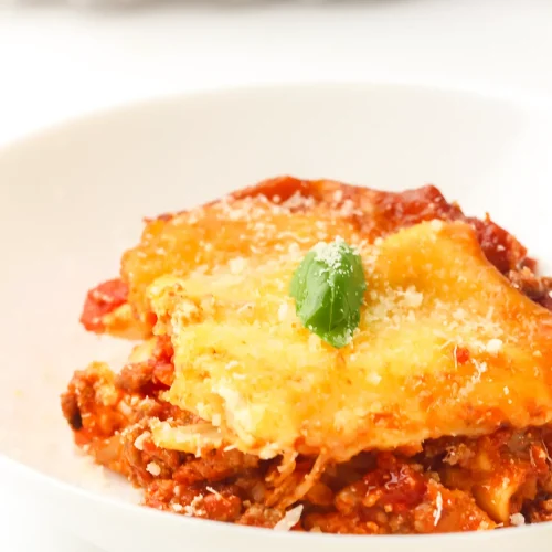 Crockpot lasagna slice in a white bowl topped with grated Parmesan and a basil leaf