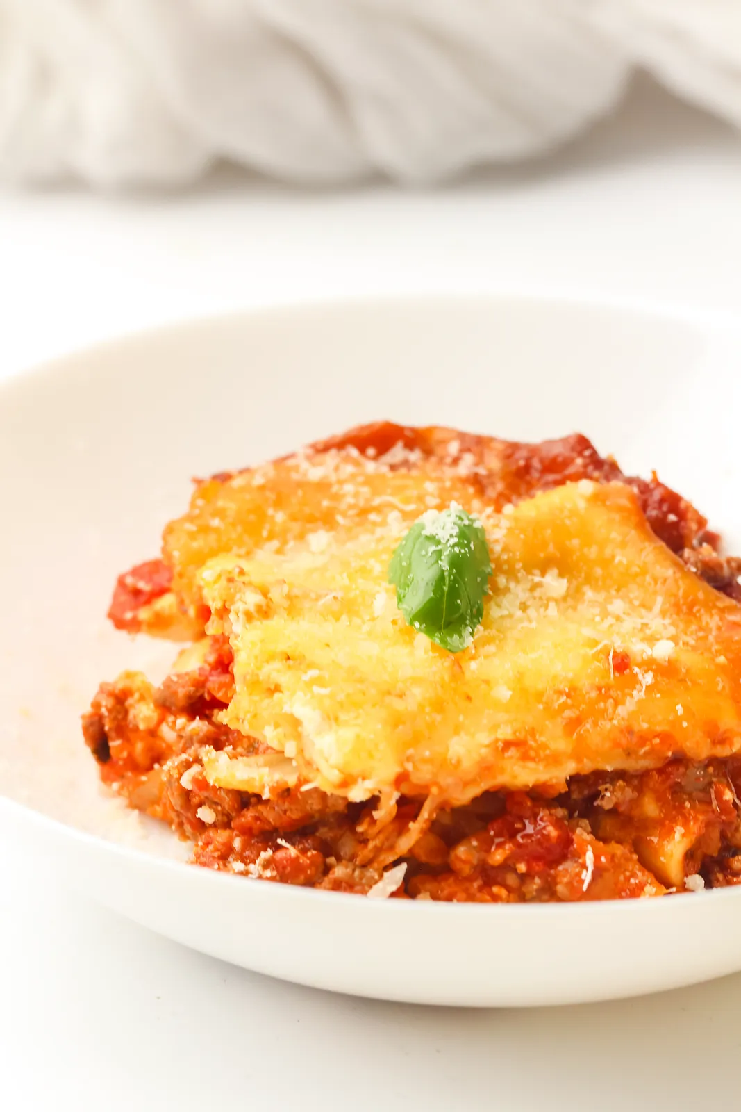 Crockpot lasagna slice in a white bowl topped with grated Parmesan and a basil leaf