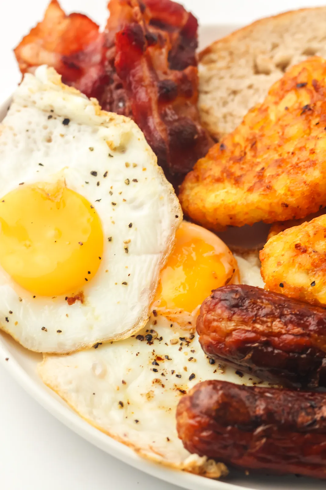 Close up of a peppered fried egg with crispy bacon, sausages and toast on a white plate