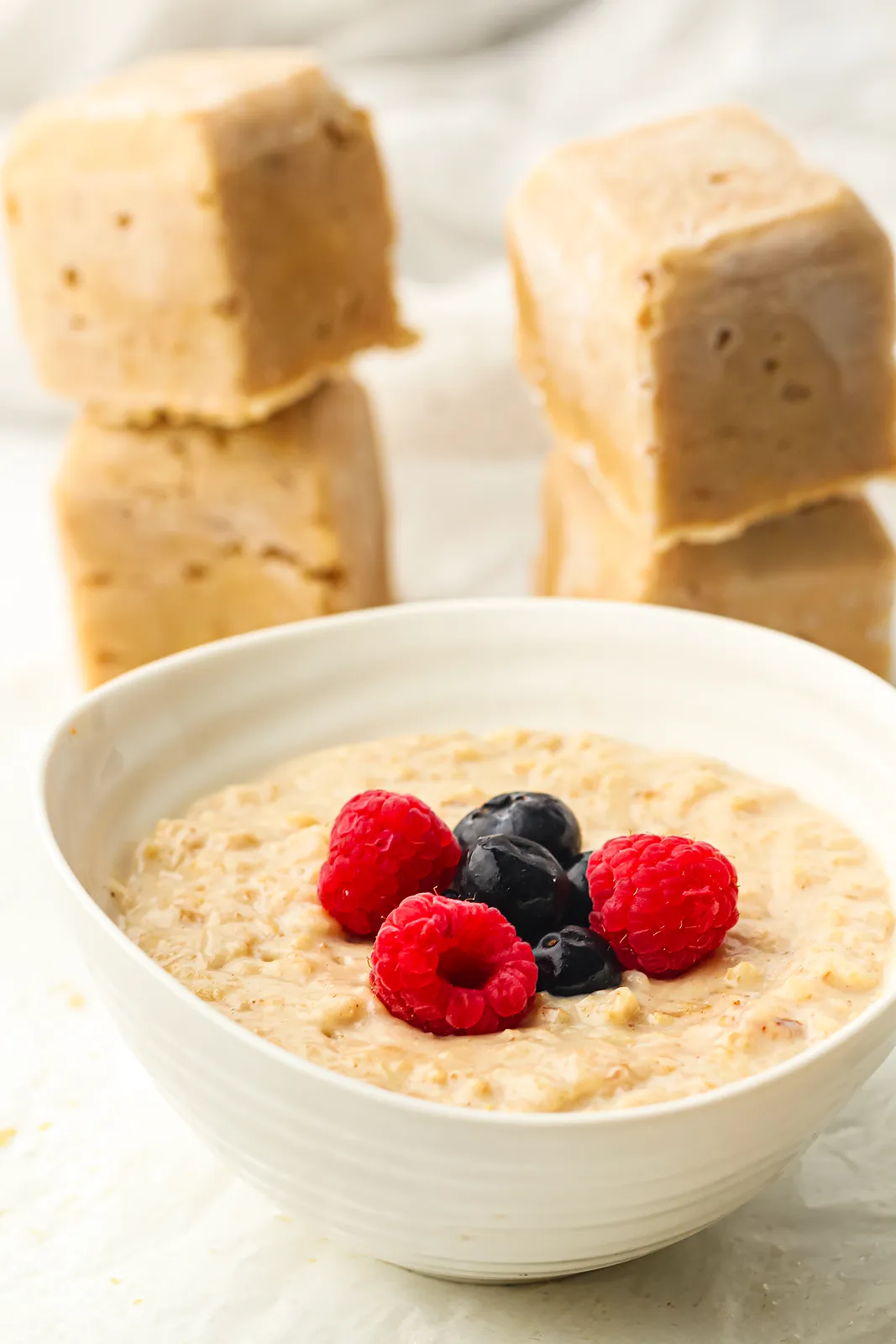 Frozen oatmeal cubes stacked beside a bowl of creamy porridge topped with raspberries and blueberries