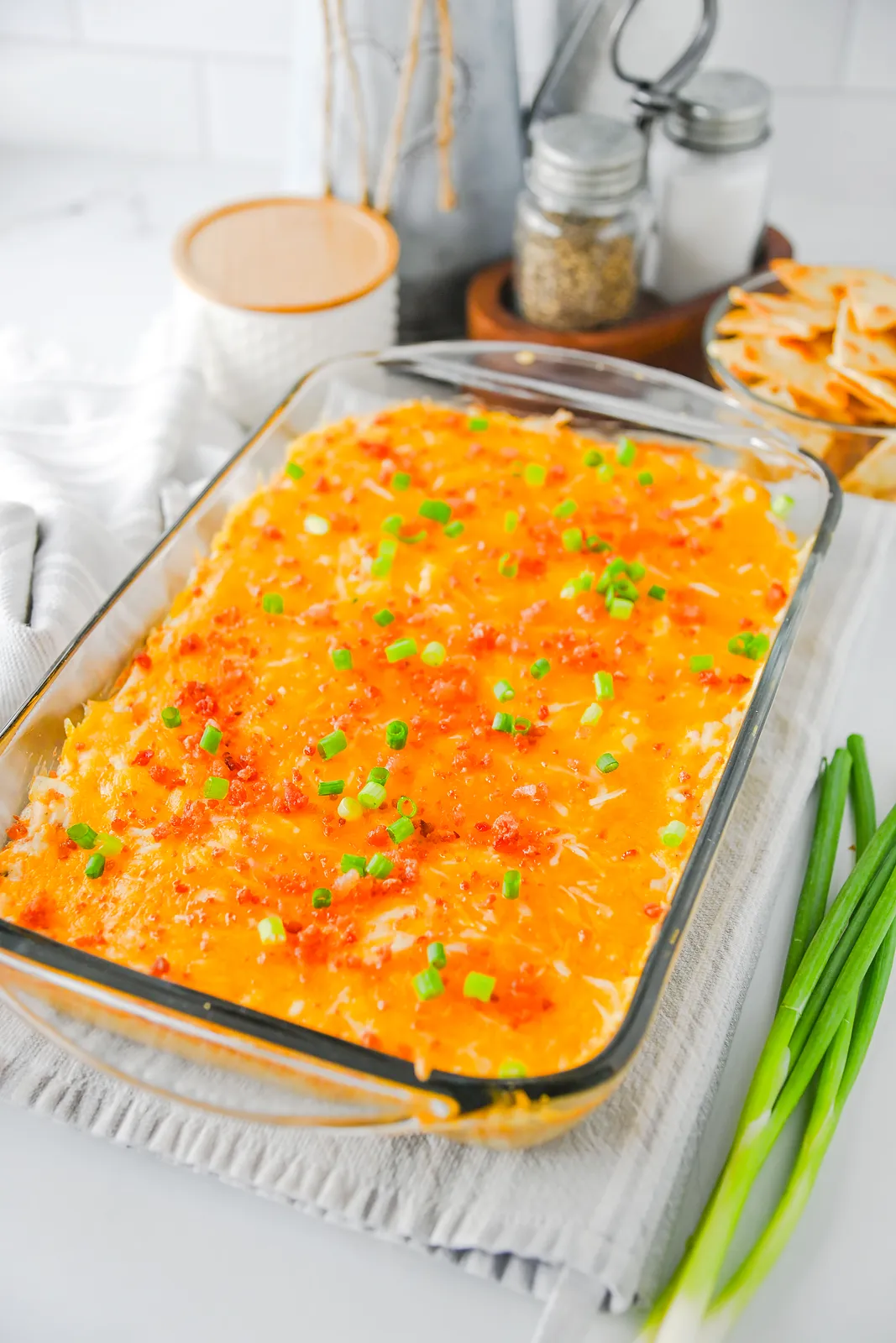 A glass baking dish of cheesy ranch and bacon appetiser with tortilla chips and spring onions on the side