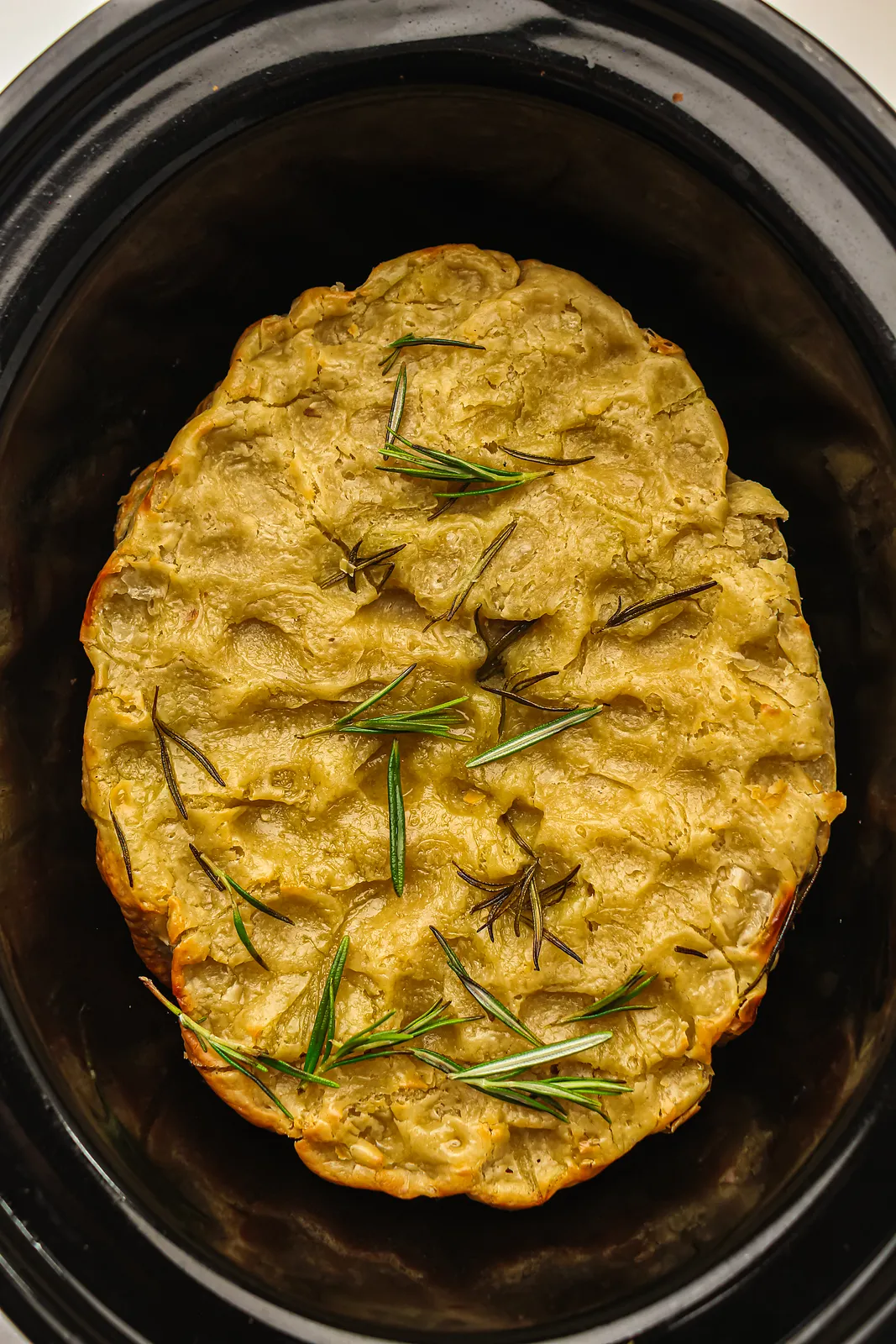 A golden, rosemary-topped loaf sitting inside a black slow cooker pot