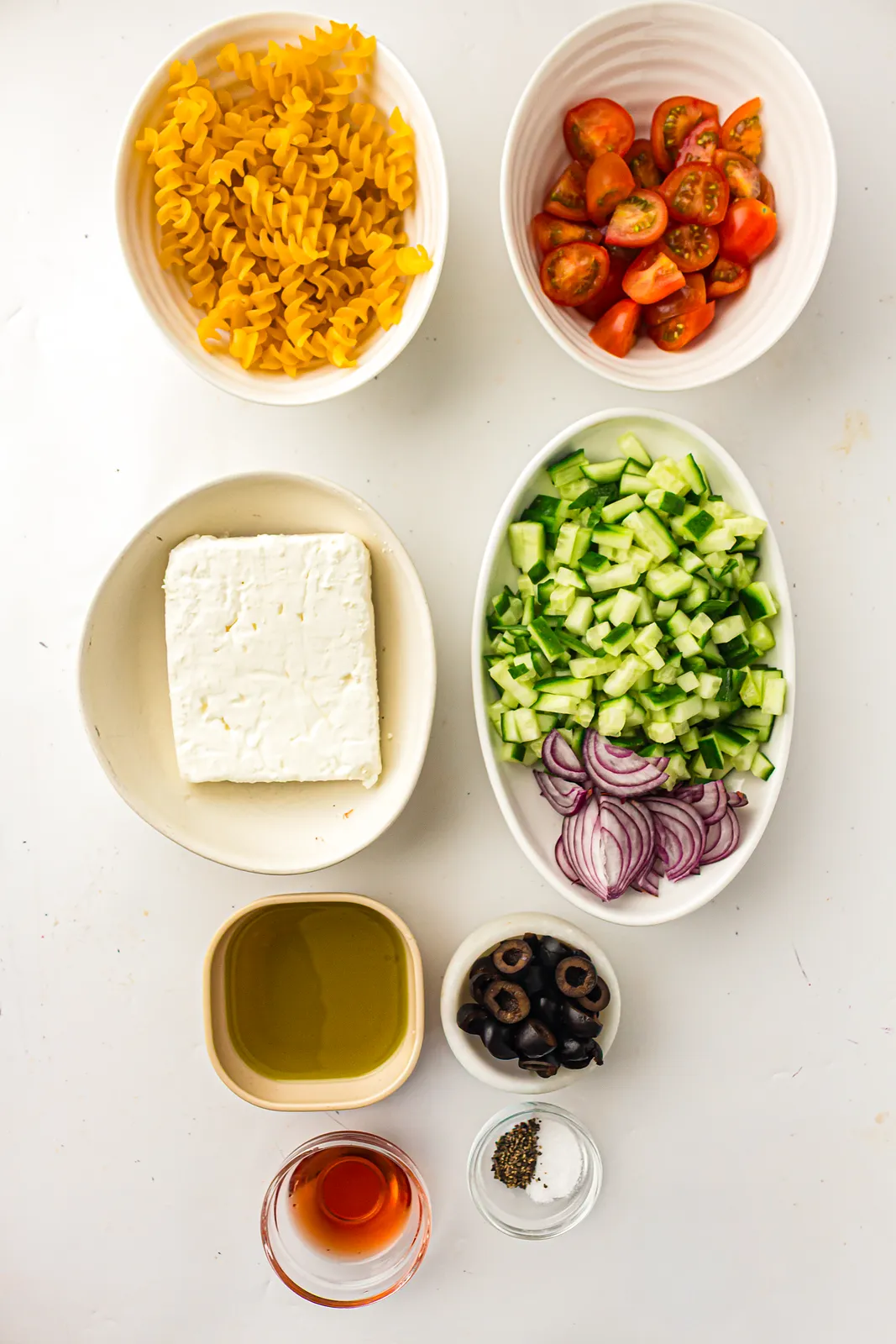 Ingredients for Greek pasta salad: rotini, cherry tomatoes, feta block, cucumber, red onion, olives, olive oil and red wine vinegar