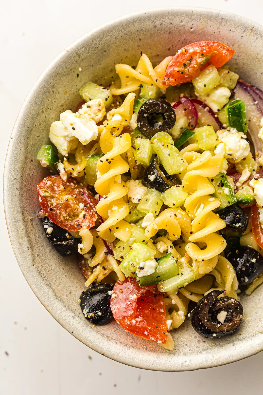 Greek pasta salad with rotini, black olives, feta, cucumber and cherry tomatoes in a speckled bowl