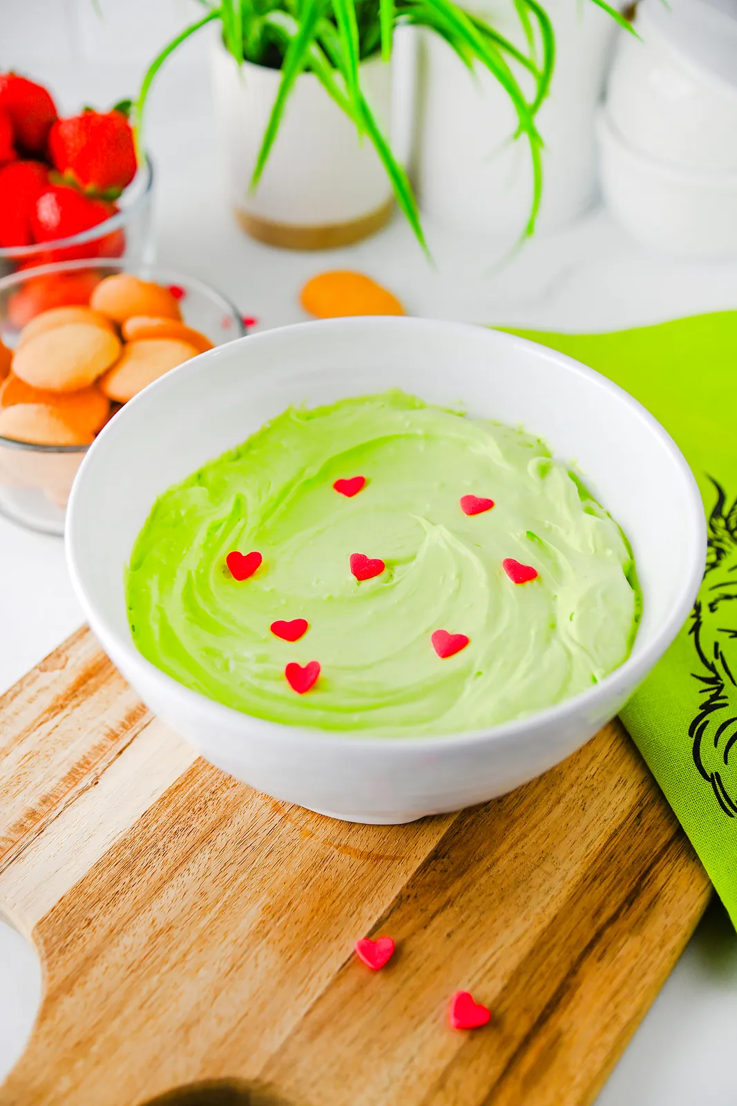 Grinch dip topped with red heart sprinkles in a white bowl on a wooden board