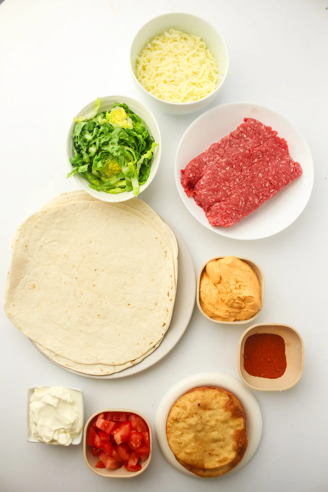 Ingredients for a homemade crunchwrap: ground beef, flour tortillas, tostadas, nacho cheese, taco seasoning, sour cream, lettuce, tomato and cheddar