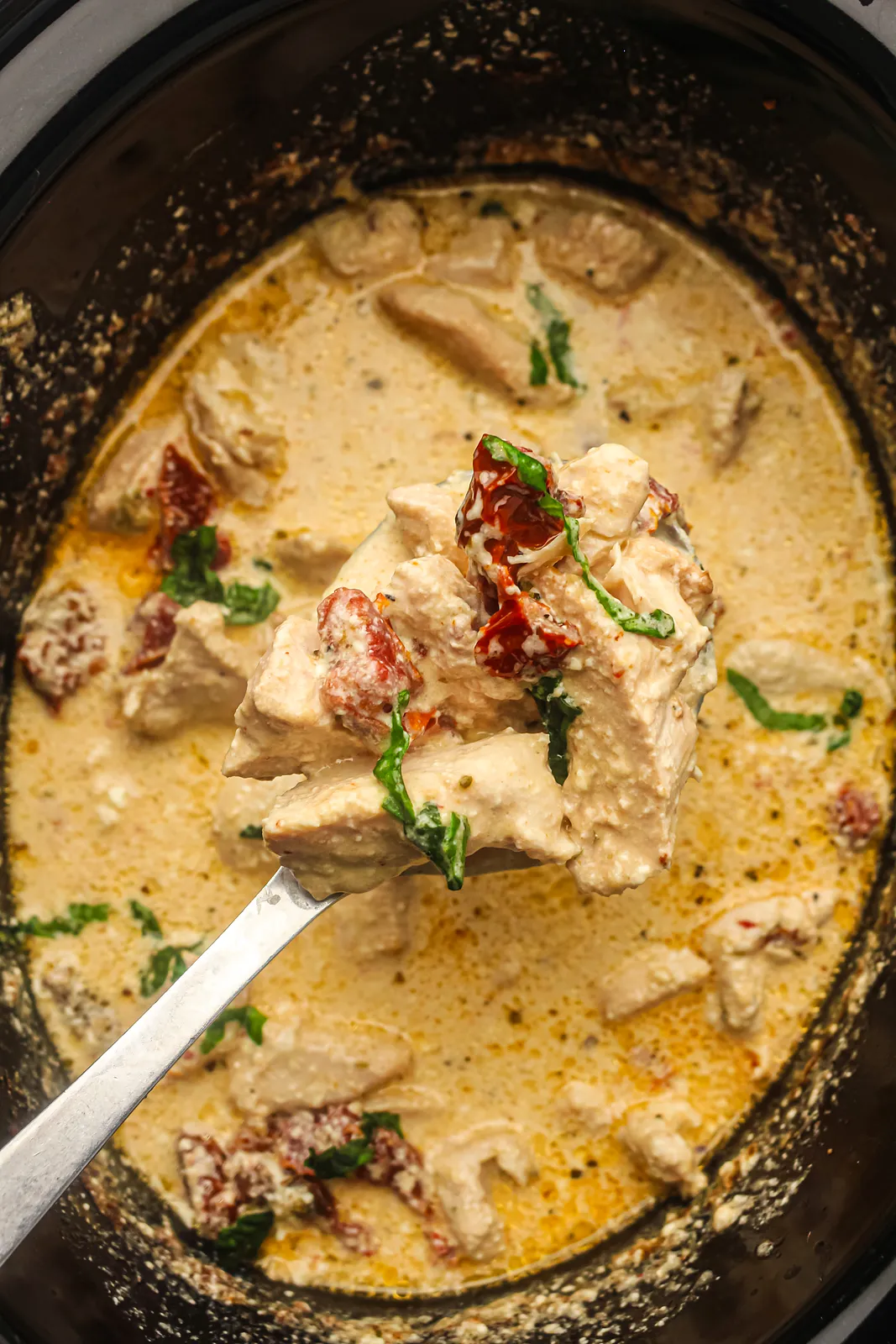 A ladle lifting creamy pieces and sun-dried tomatoes from the black slow cooker pot