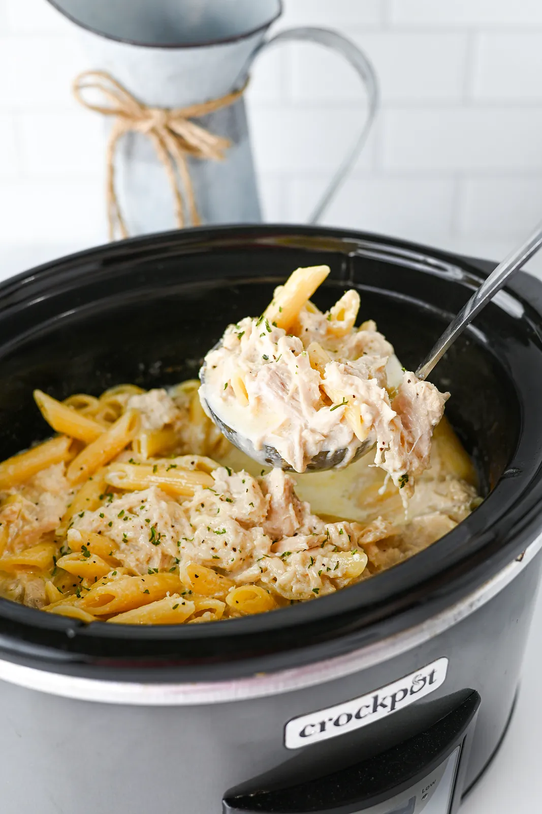 A ladle lifting creamy penne and shredded meat out of the slow cooker