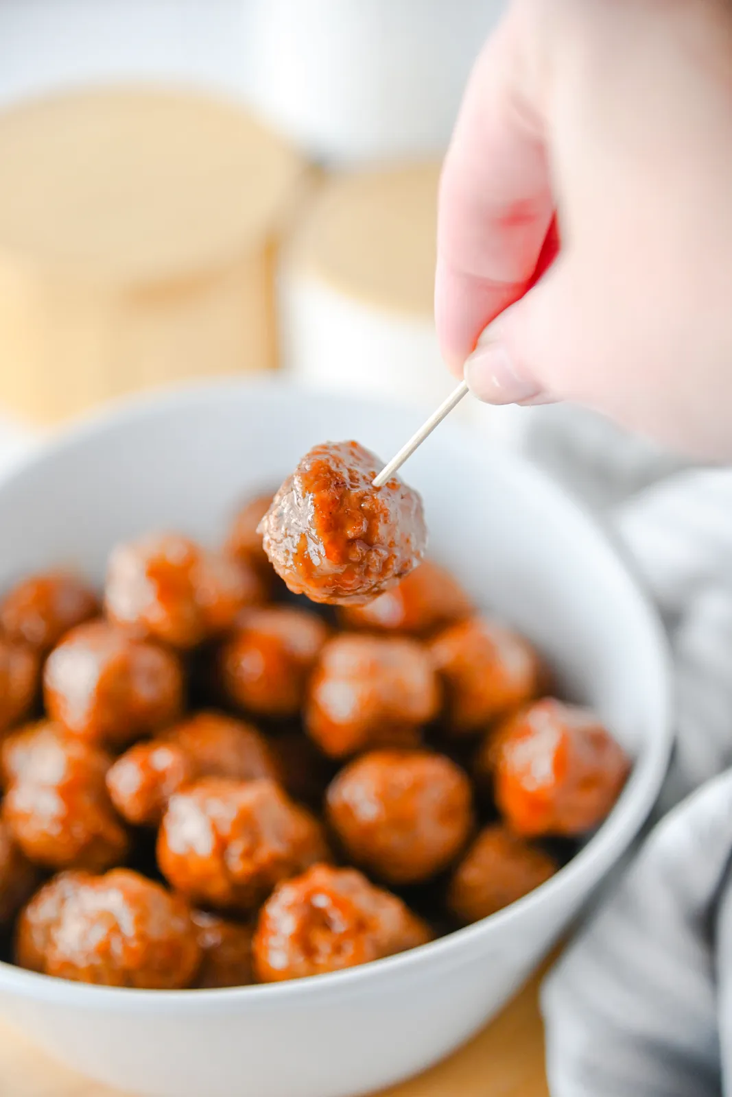A hand lifting a sticky glazed meatball on a toothpick from a white bowl