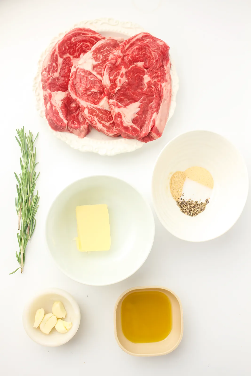 Ingredients for ribeye on Blackstone: raw marbled steaks, rosemary, butter, garlic, olive oil and a bowl of seasoning