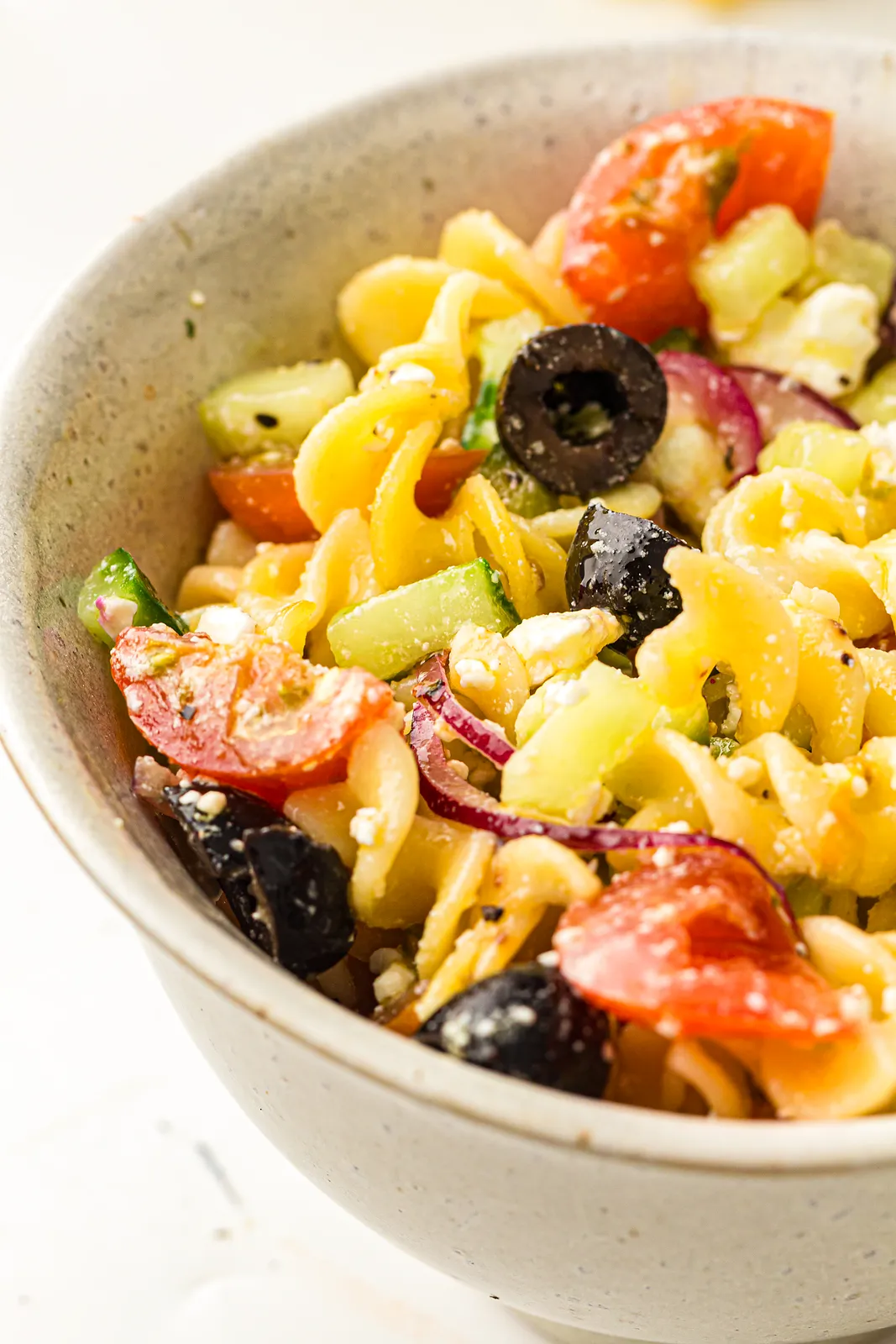 Rotini spirals with black olive slices, halved cherry tomatoes and cucumber in a cream bowl