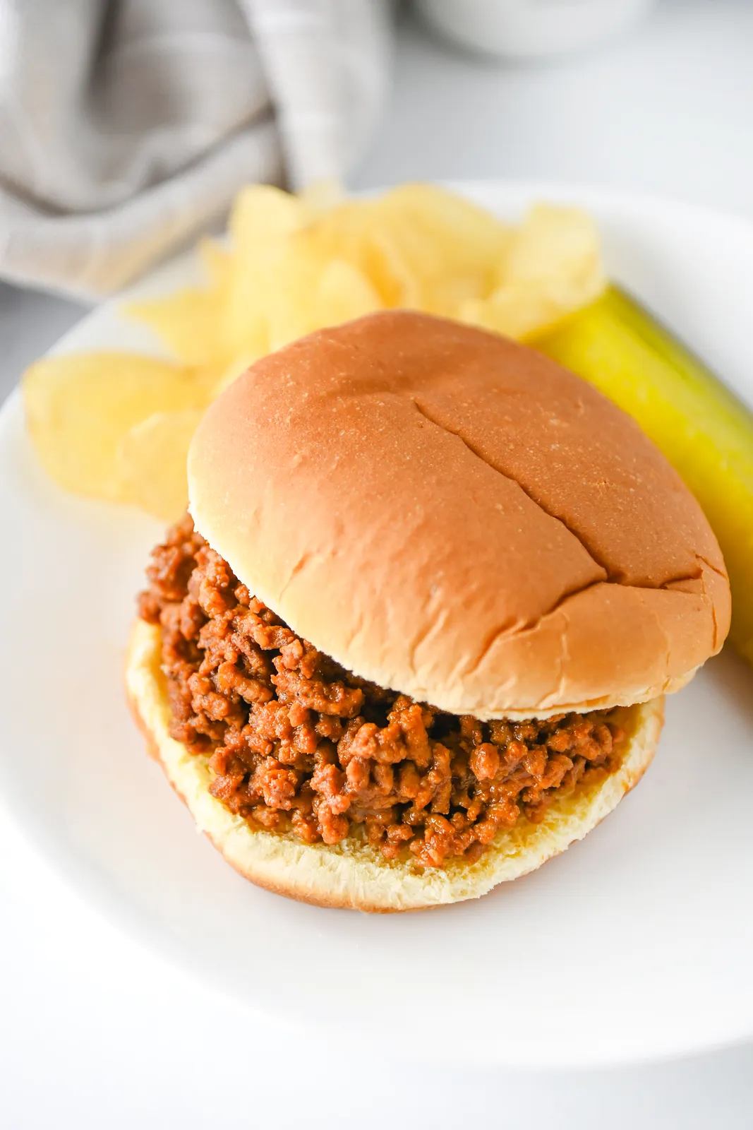 A saucy beef bun on a white plate with crisps and a dill pickle spear