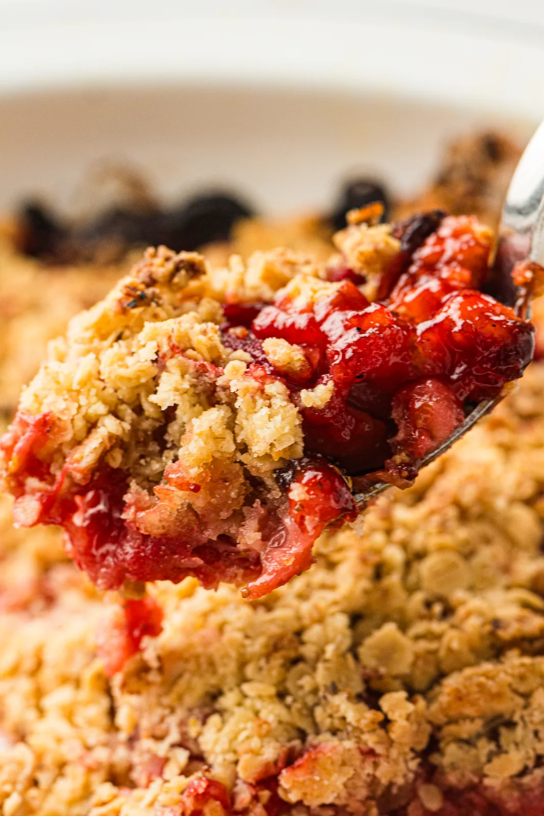 A spoon lifting a jammy portion of red berries and crisp oat topping from the dish