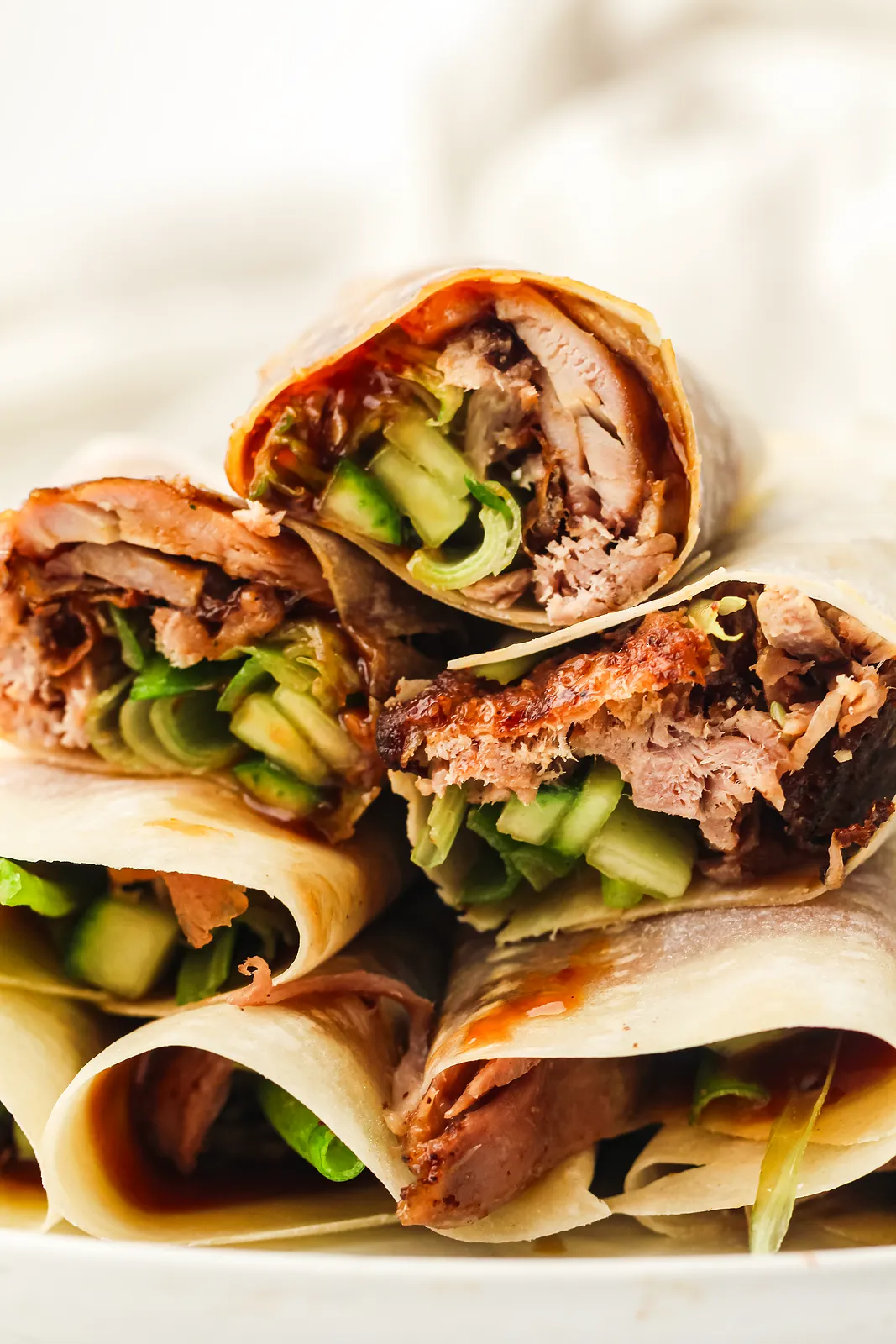 A stack of rolled wraps showing crispy meat, hoisin and green cucumber inside