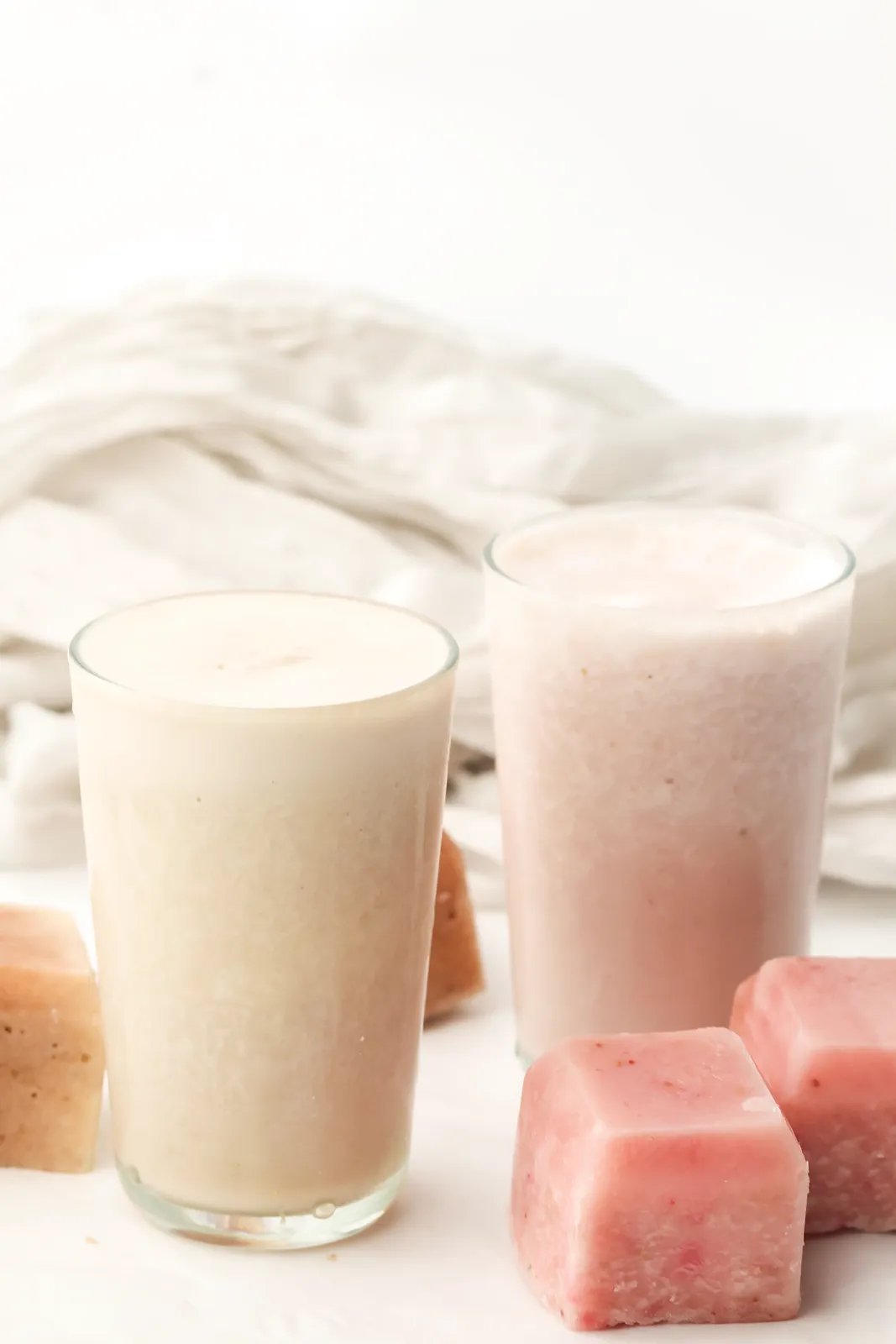 Two glasses of thick blended drink, one pale pink and one creamy, with pink frozen blocks beside them