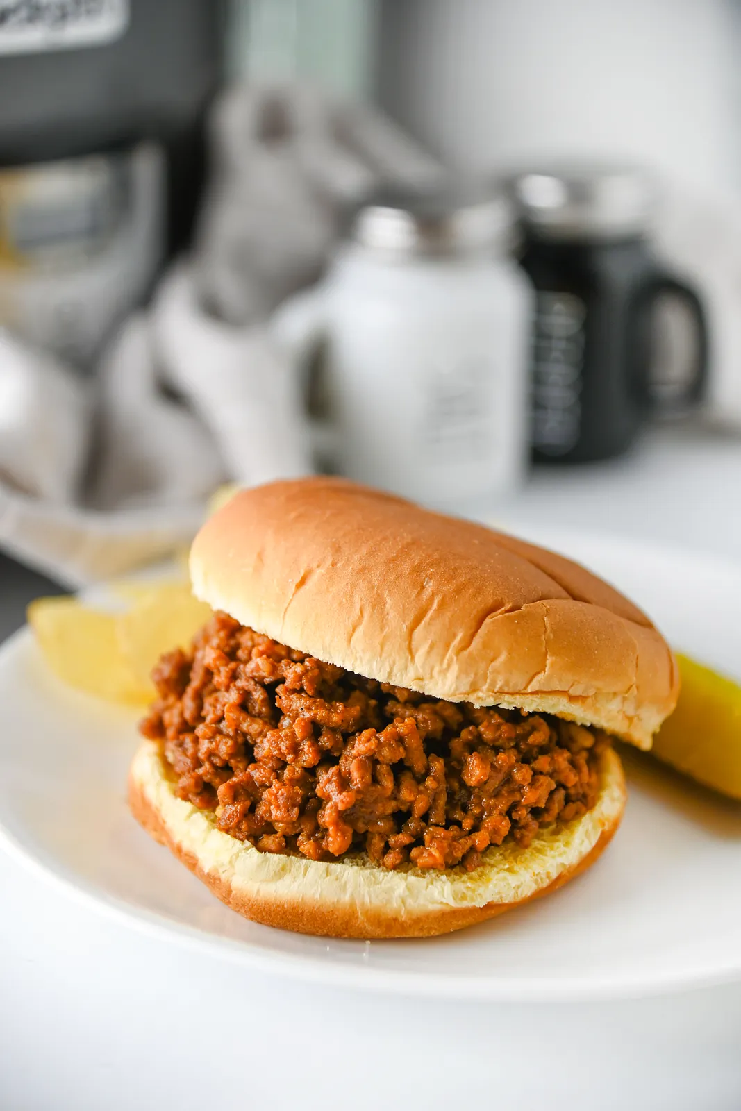 A tangy beef sandwich on a white plate with a pickle on the side