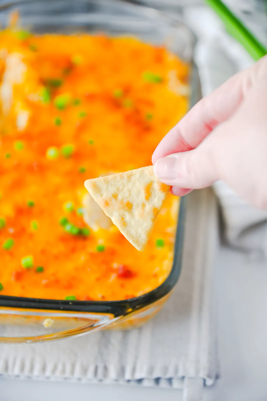 A hand pushing a tortilla chip into the hot, cheesy baking dish