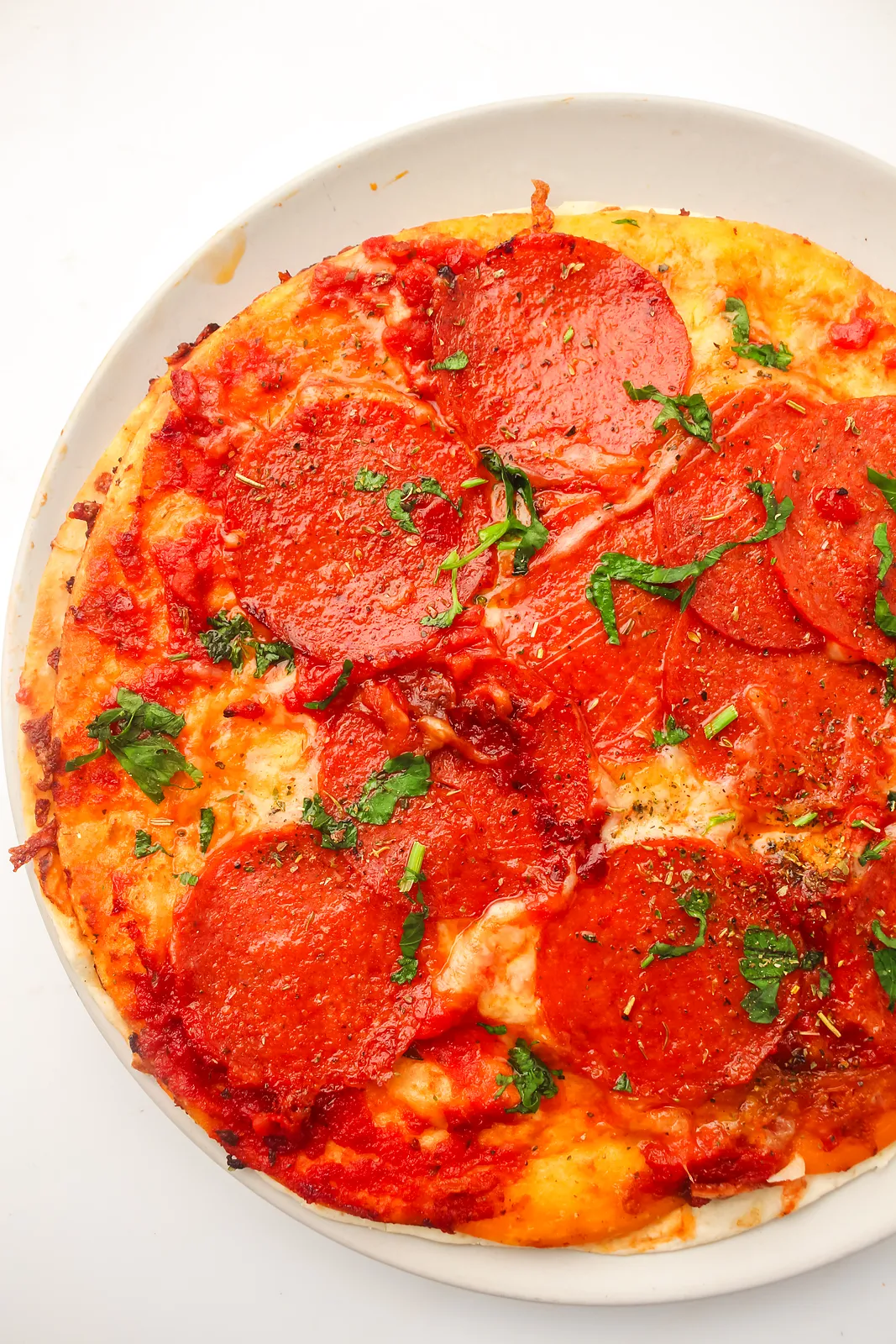 Tortilla pizza with crispy pepperoni, melted mozzarella and herbs on a white plate