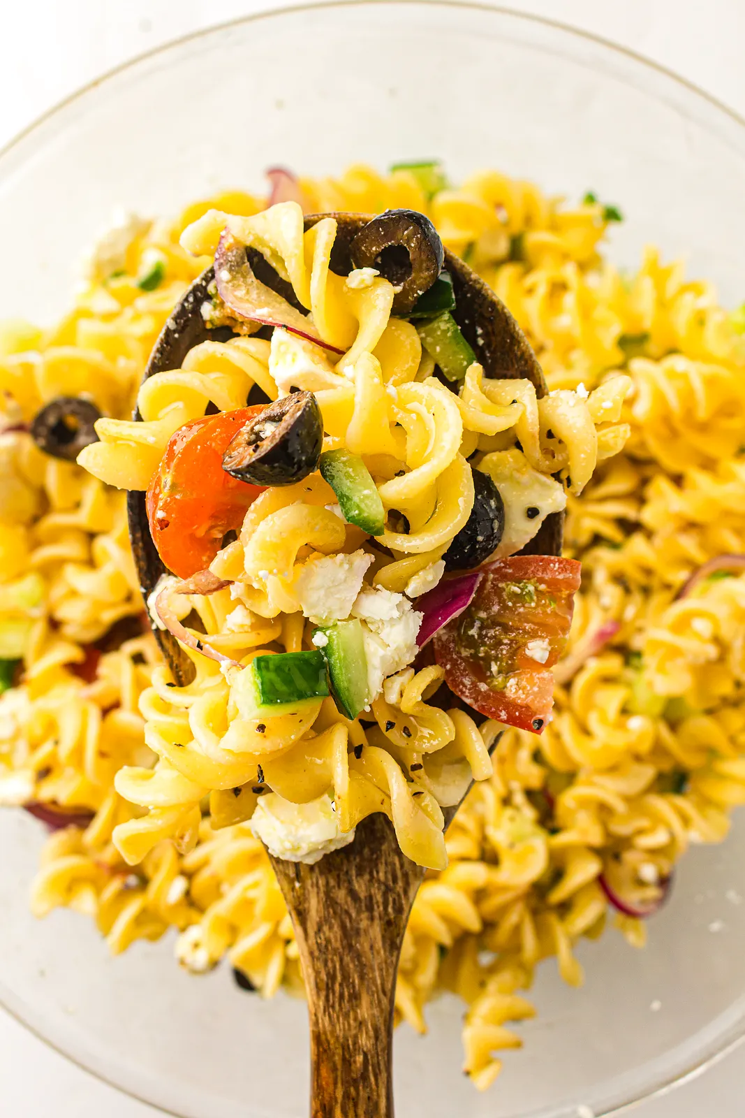 A wooden spoon lifting glossy rotini with tomato, olive and cucumber from a glass bowl